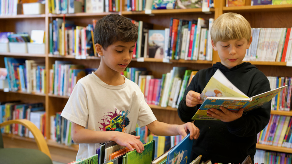 Två barn som koncentrerat bläddrar i och bland böcker i ett bibliotek.