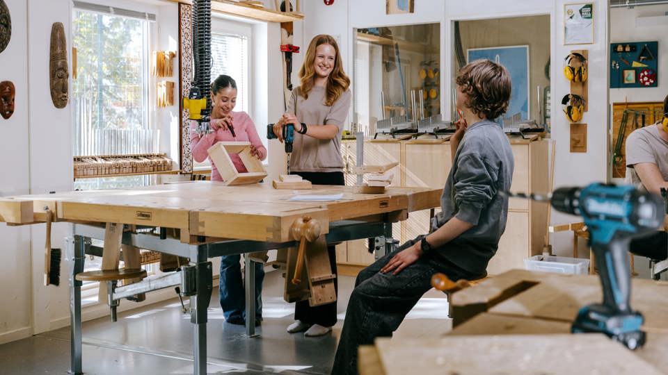 Elever arbetar med träslöjd vid ett stort arbetsbord och använder verktyg i en ljus slöjdsal.
