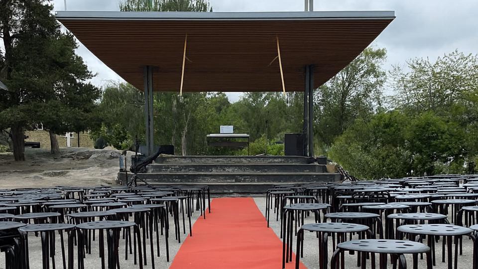 Svarta pallar och en röd matta är utplacerades framför en scen. Lövträd syns bakom och intill scenen.
