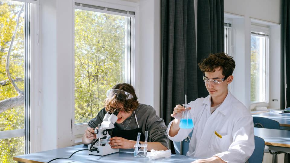 Två elever utför naturvetenskapliga experiment i ett klassrum. En tittar i ett mikroskop, medan den andra håller en mätkolv med blå vätska.