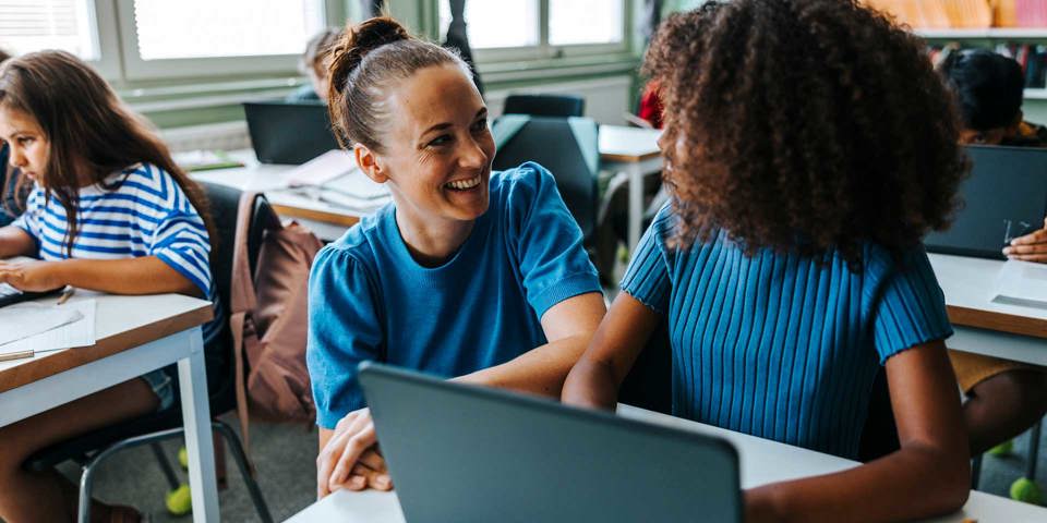 Kvinnlig lärare och elev vid skolbänk ler mot varandra 