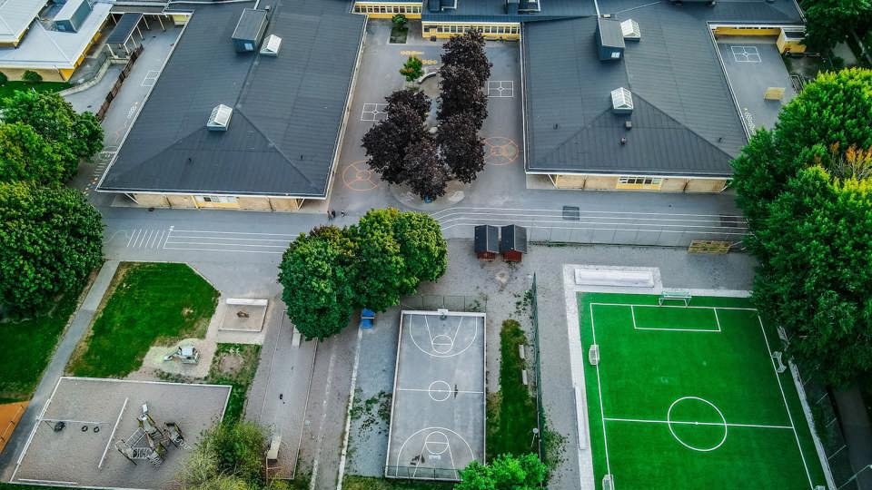 En flygbild över en skolas gård med byggnader, träd, lekplatser samt basket- och fotbollsplaner.