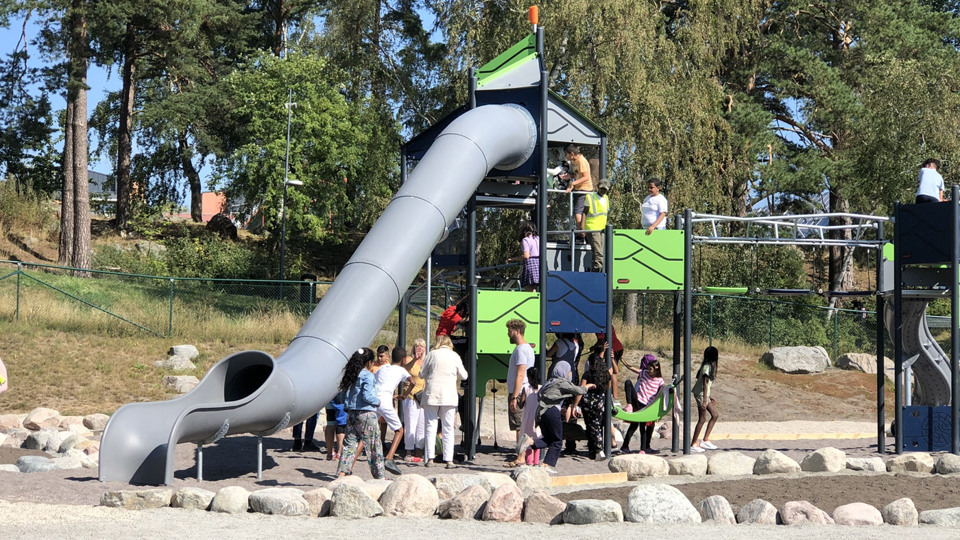 Barn som klättrar i en stor klätterställning med rutschkana på en skolgård. I bakgrunden syns en träddunge och hus.